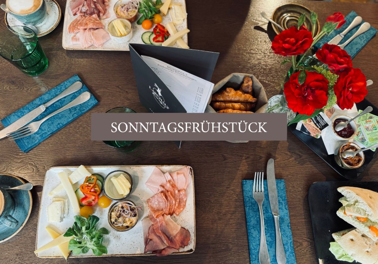 Ein Tisch zeigt ein Sonntagsfrühstücksmenü mit Fleisch, Käse, Gemüse und Brot. Blumen und Besteck sind ebenfalls vorhanden.