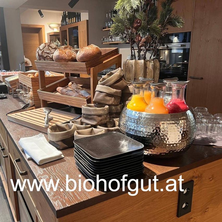 Ein Frühstücksbuffet mit Brot, Getränken und einer Website-Adresse. Verschiedene Brotsorten sind auf einem Holztablett. Eine Pflanze in einem Vasen und ein Krug mit Saft sind in der Nähe.