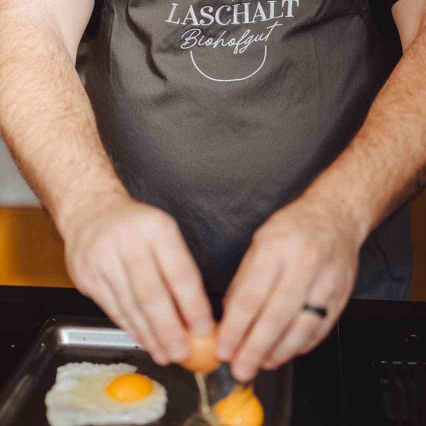 Eine Person kocht Eier auf einem Tablett in einer Küche. Sie trägt ein schwarzes Apron mit den Worten Laschalt Biohofgut.