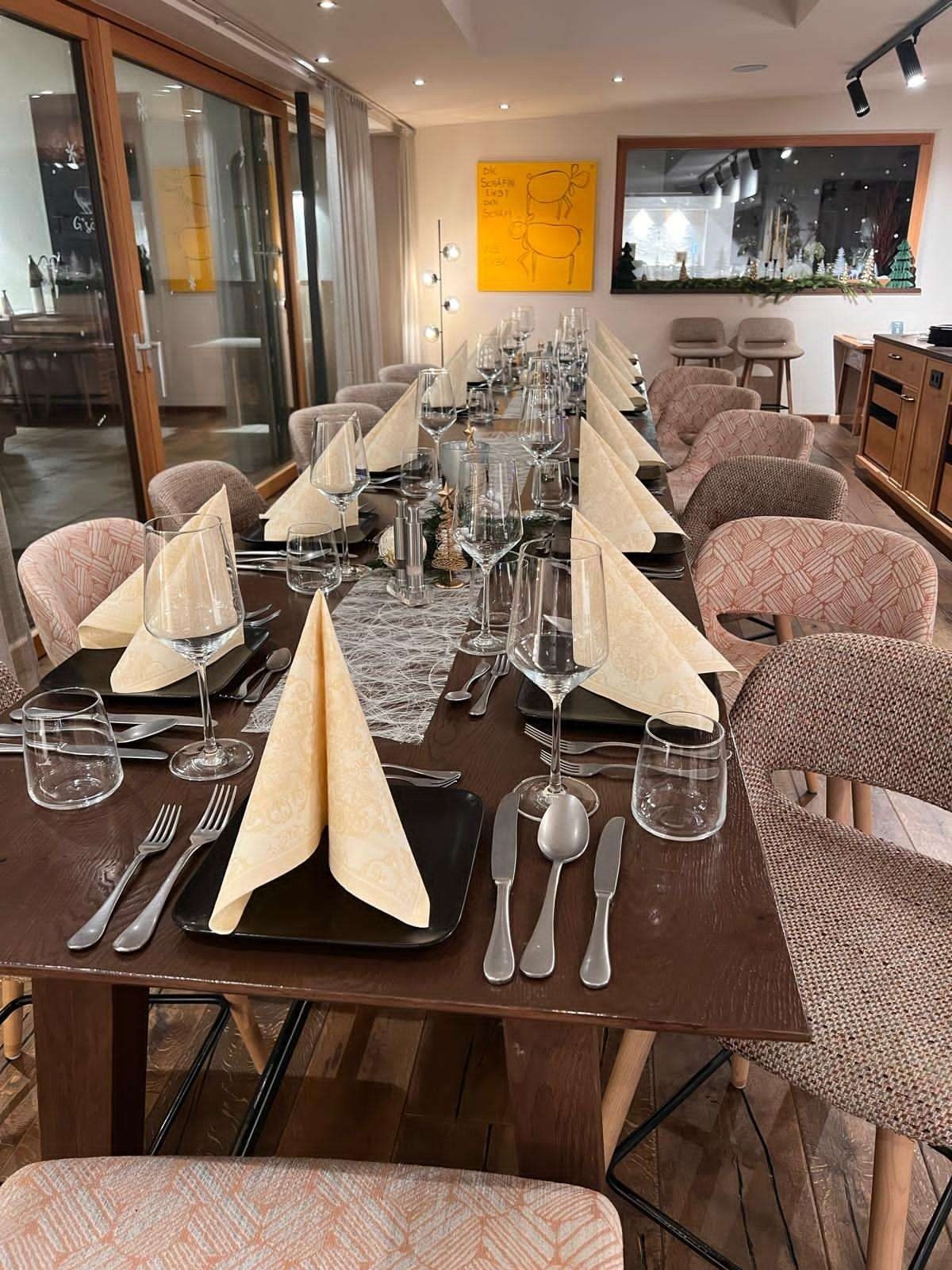 Ein langer Esstisch mit Tischdecken, Gläsern, gefalteten Servietten, Besteck und Stühlen in einer Restaurantumgebung mit festlicher Kulisse.
