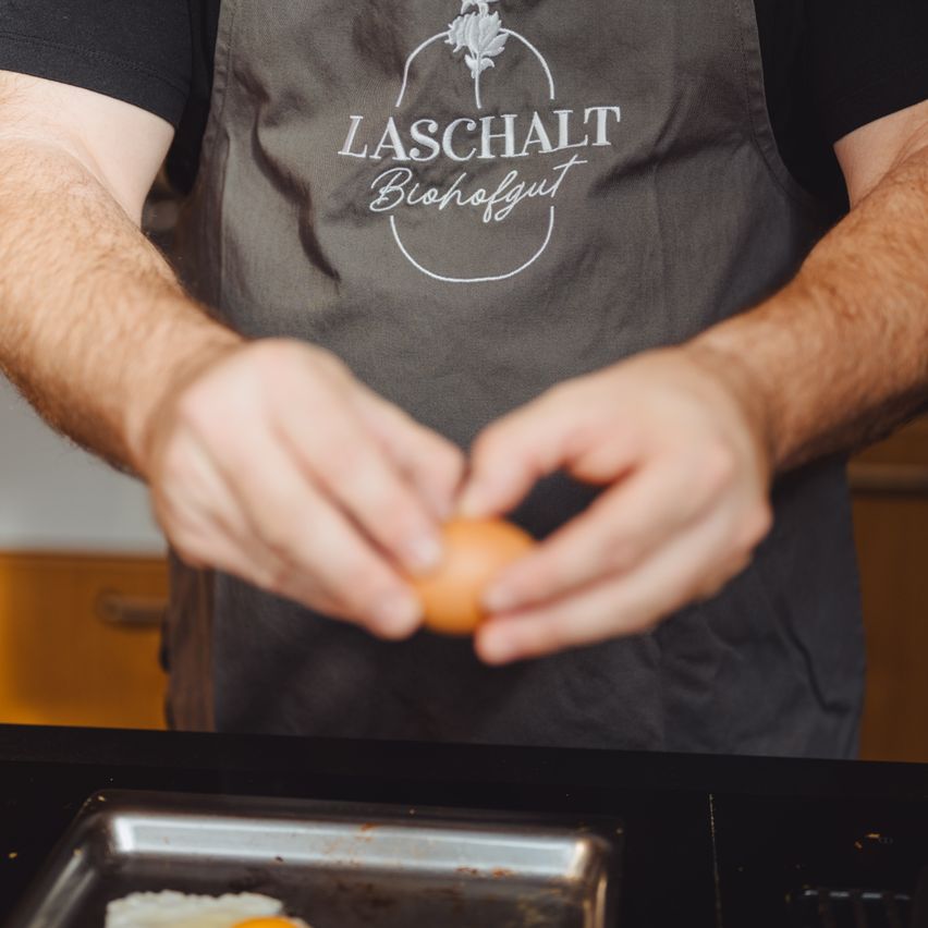 Eine Person in einer Laschalt-Schürze knackt ein Ei auf einem Tablett in der Nähe eines Grills.