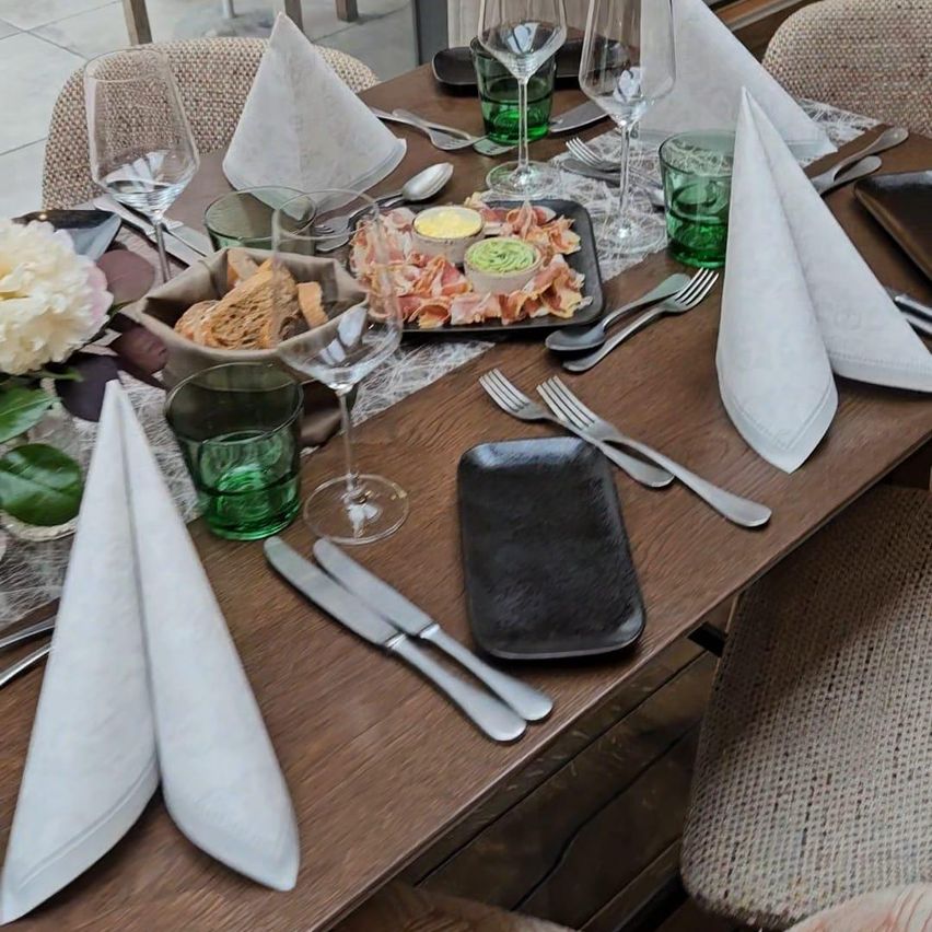 A wooden table with neatly set place settings, including folded napkins, glasses, and a dish of meats and cheeses. A white flower adds a touch of color.