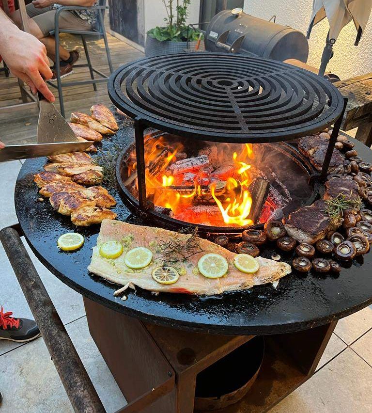 A person is grilling meat and fish on a black outdoor grill with fire and herbs on a tiled floor.