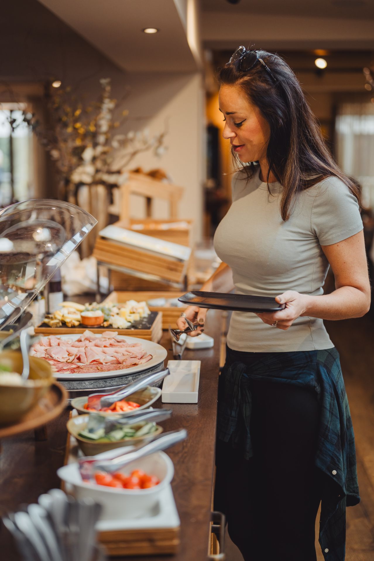 A woman wearing a gray shirt and black pants is standing behind a table with various food items on it. She is holding a plate and appears to be serving.
