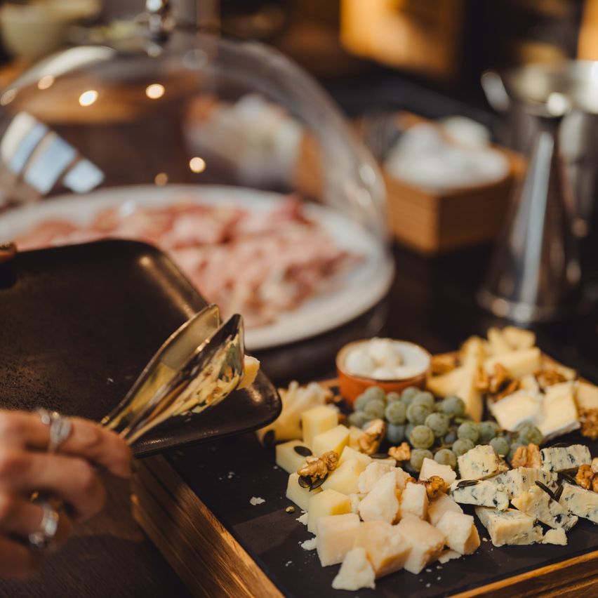 A cheese platter with various cheeses, grapes, and nuts is presented. Nearby, a tong is used to pick up cheese from a tray. Another dish and a covered glass dome are visible in the background.