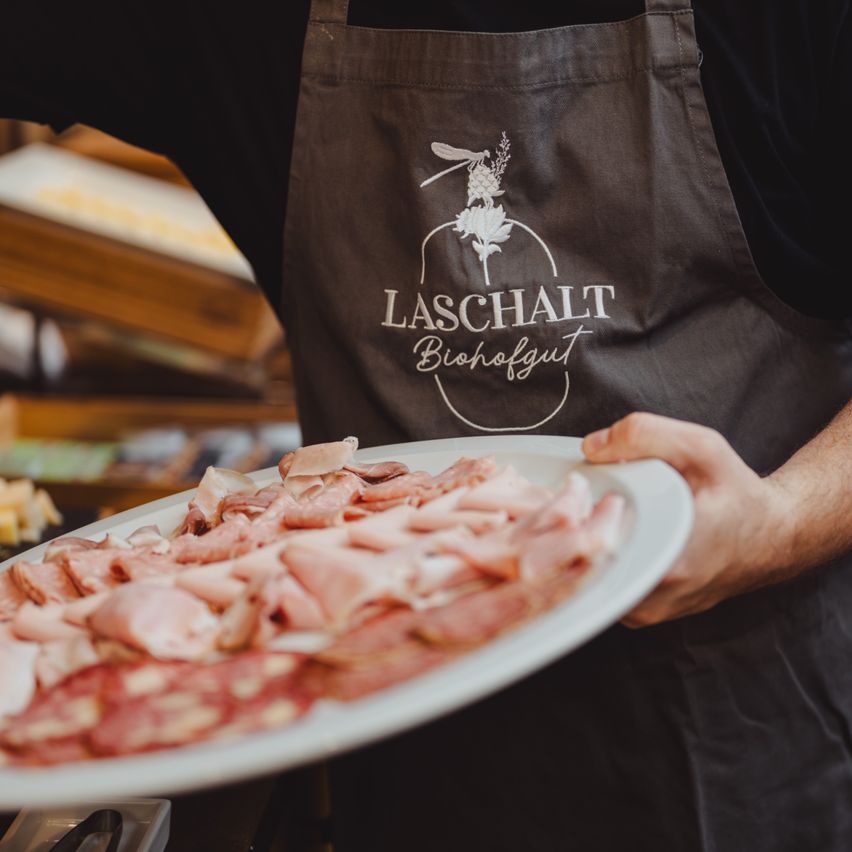 A person in a 'LASCHALT Biohofgut' apron holds a large white plate filled with sliced meats. The background features a wooden counter and assorted foods.
