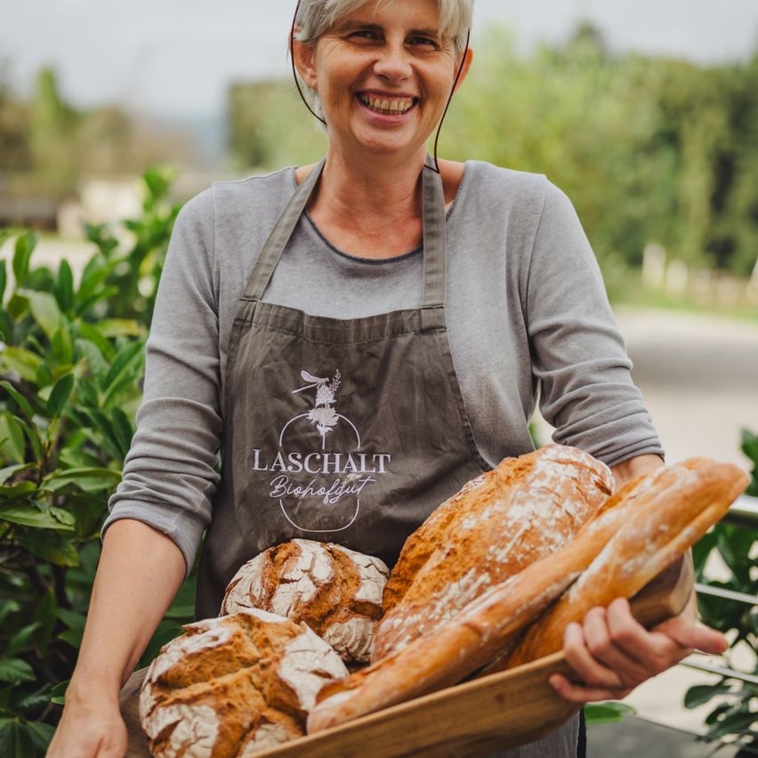 A woman wearing an apron with the logo 'LASCHALT Biohofgut' holds a tray with various types of bread in front of a garden background.