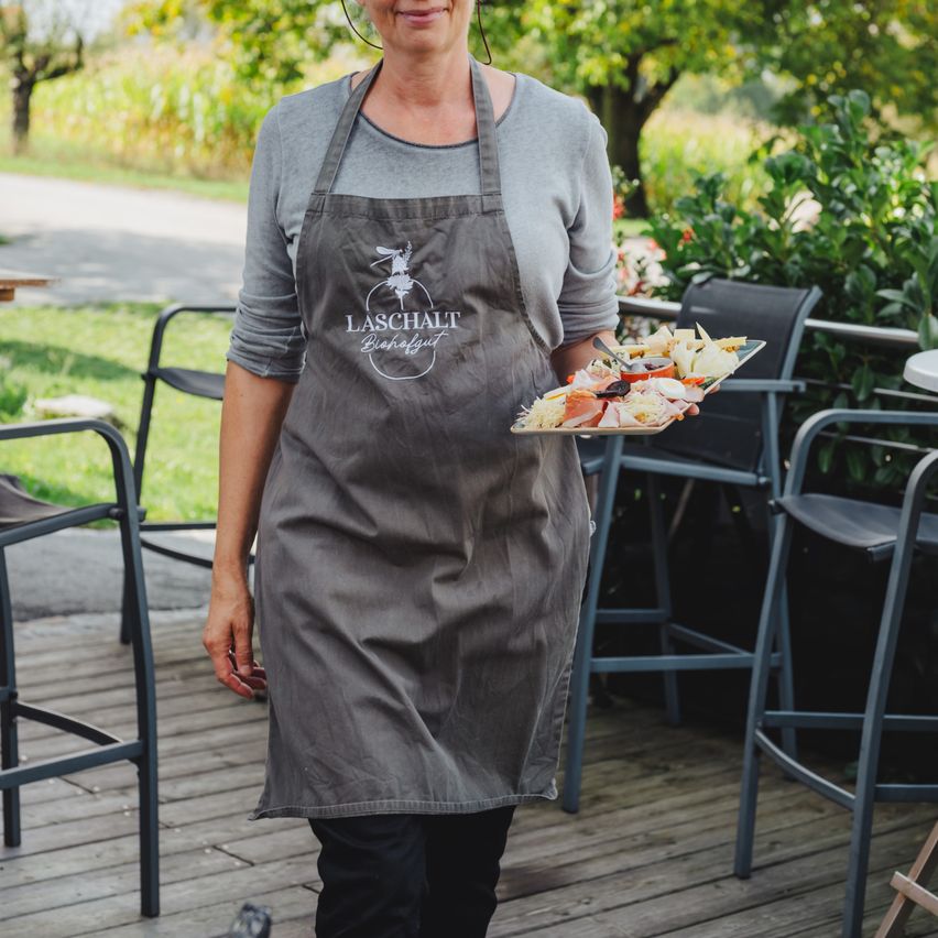 Eine Frau in einer Schürze hält einen Tablett mit Essen auf einer Holzterrasse mit Stühlen und Pflanzen im Hintergrund.