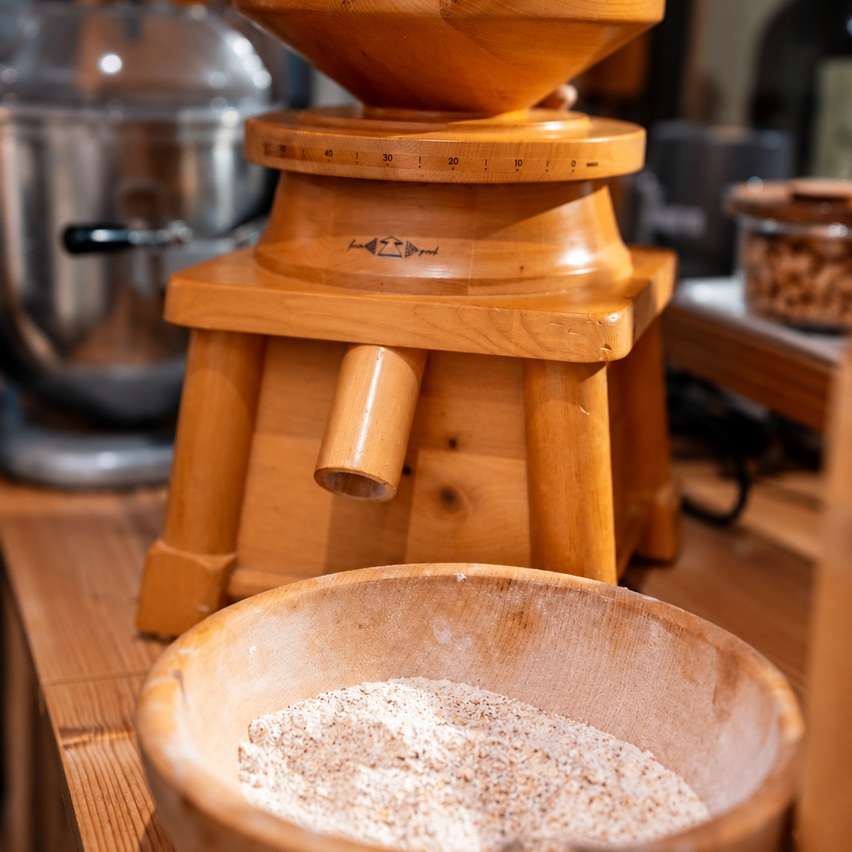 A wooden grain mill with a wooden bowl filled with grain in front of a stainless steel mixer.