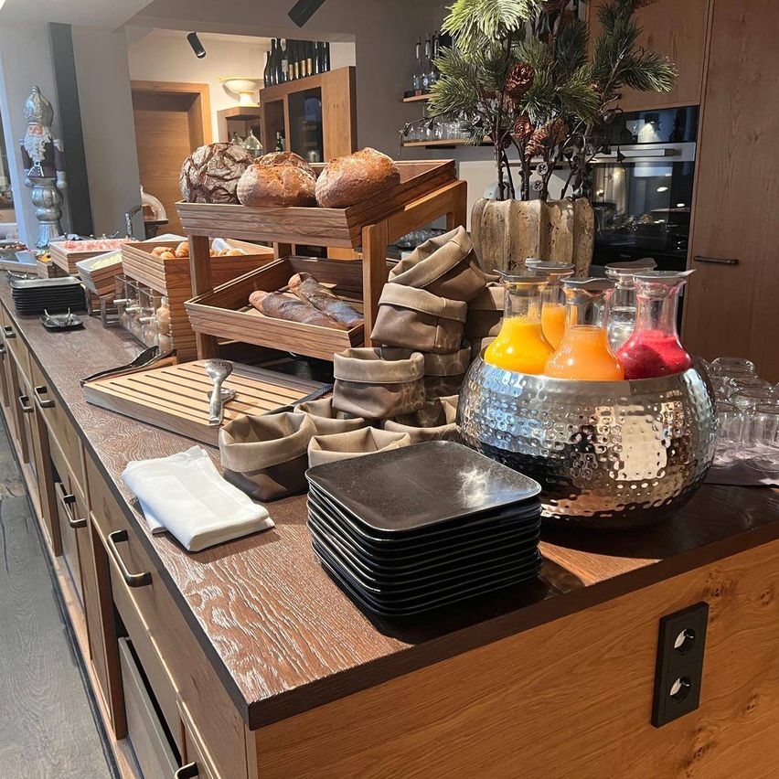 A breakfast buffet with various breads, fresh juices, and a decorative plant in a wooden setting.