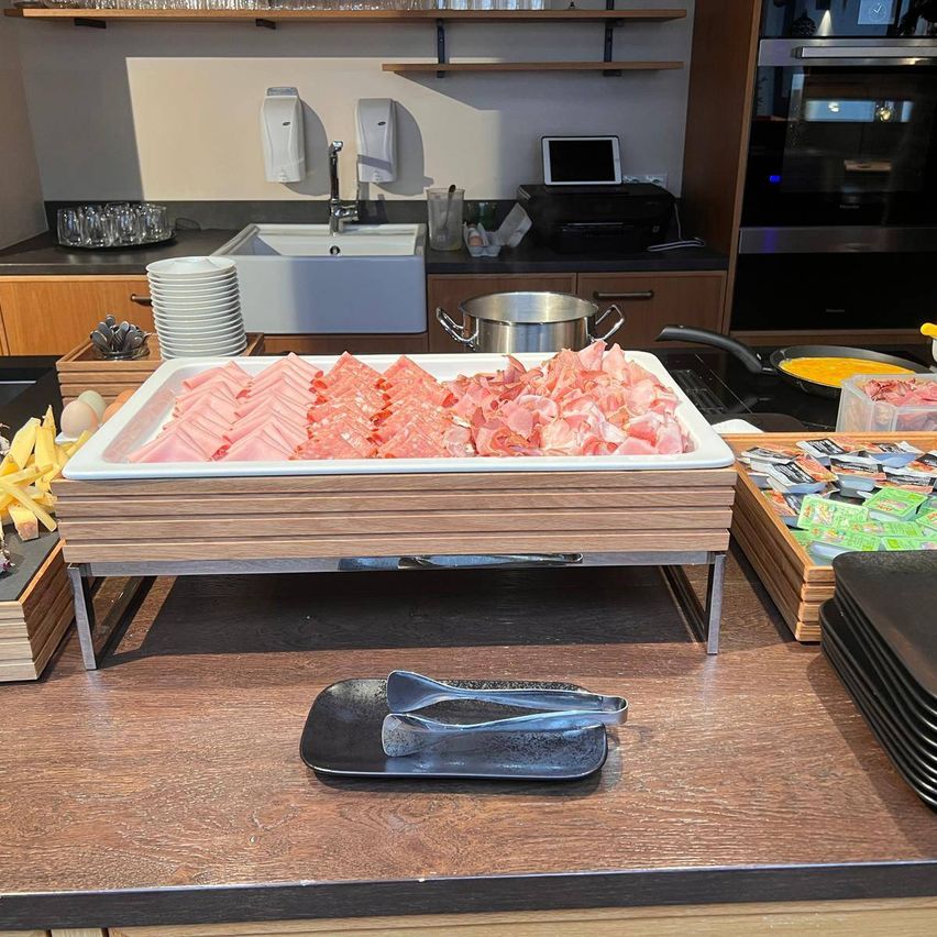 A kitchen with a wooden countertop, a tray of sliced meat, and utensils. Behind it, a sink, a stove, and a refrigerator.