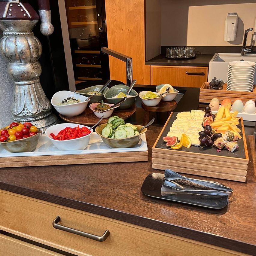 A wooden kitchen counter with a variety of food items on it. There are bowls of vegetables, cheese, and fruit. There is also a tray with eggs and a bottle of hand sanitizer.