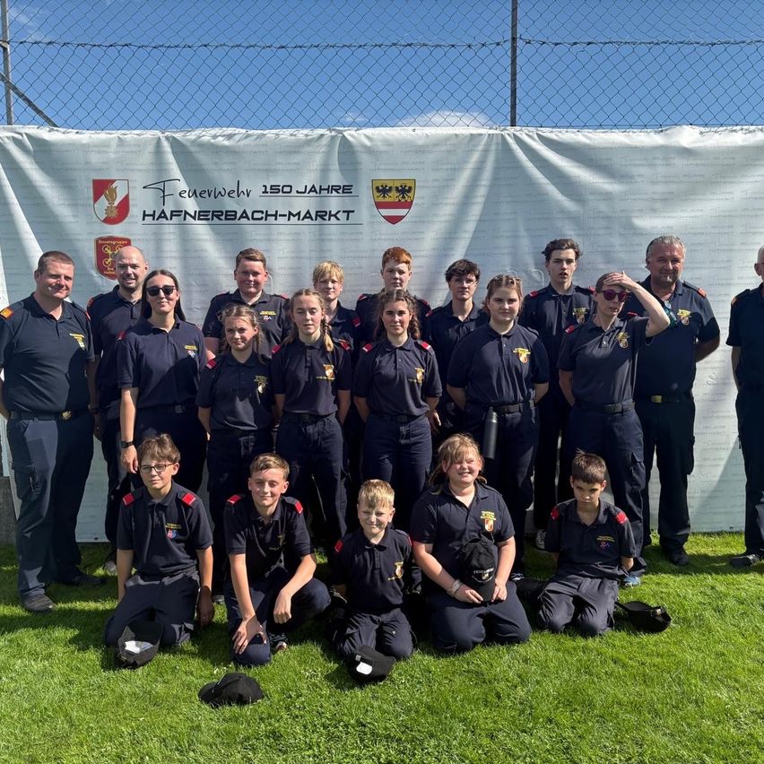 Eine Gruppe von Feuerwehrleuten in Uniform posiert für ein Foto vor einem Banner, das 150 Jahre Hafnerbach-Markt feiert. Sie lächeln und stehen oder knien auf dem Rasen.