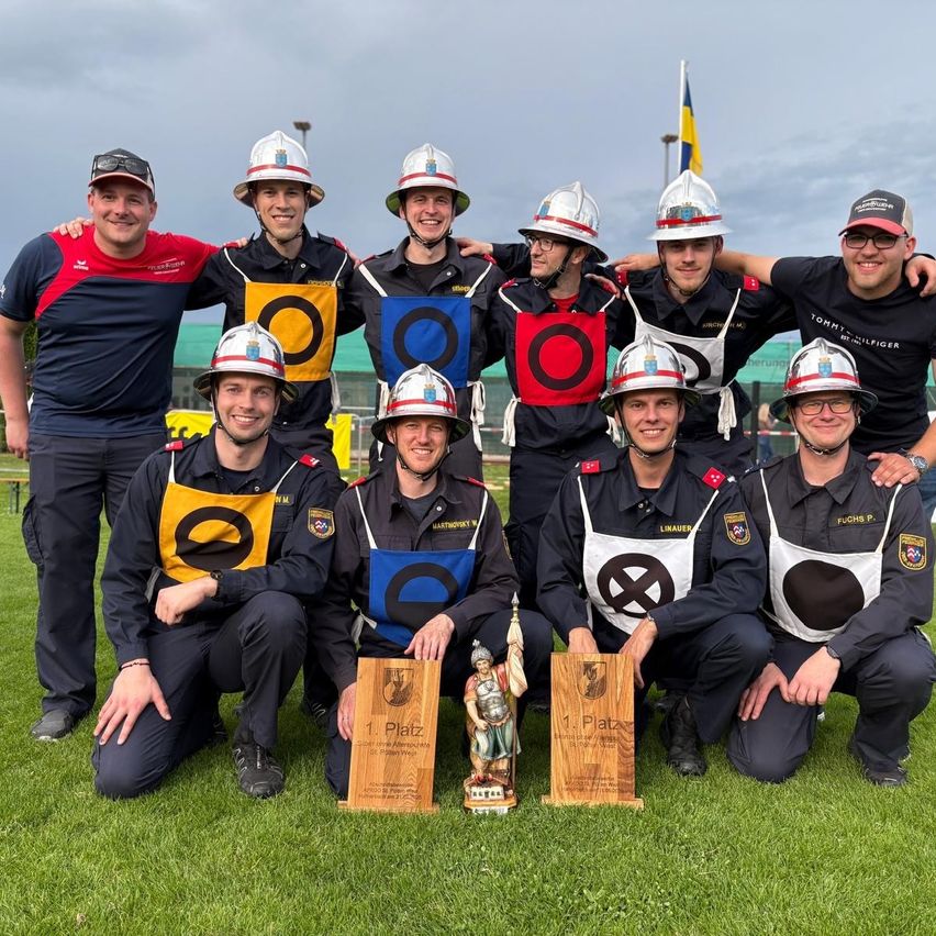 Eine Gruppe von Feuerwehrleuten posiert für ein Foto mit zwei Trophäen. Sie tragen alle Uniformen mit Helmen. Sie lächeln und posieren für die Kamera. Die Trophäen haben den Text '1. Platz' darauf. Sie stehen auf einem Rasen. Hinter ihnen ist eine verschwommene Ansicht einer Flagge und eines Gebäudes.