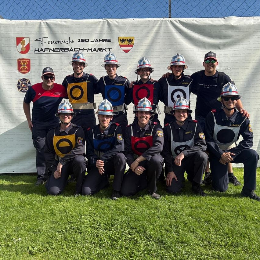 Eine Gruppe Feuerwehrleute in Uniform posiert für ein Foto vor einem weißen Hintergrund mit Logo und Text. Sie lächeln und tragen Helme und Brillen.