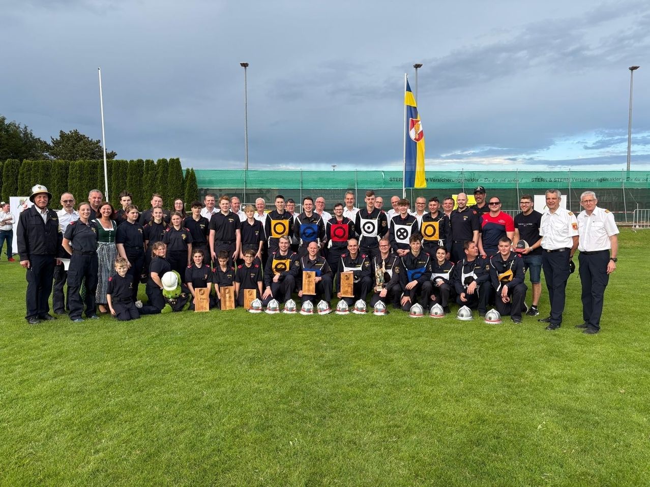 Eine Gruppe von Menschen, wahrscheinlich ein Sportteam, posiert für ein Foto auf einem Feld. Einige tragen Uniformen mit Abzeichen. Sie halten Pokale, und es gibt Stangen mit Flaggen.