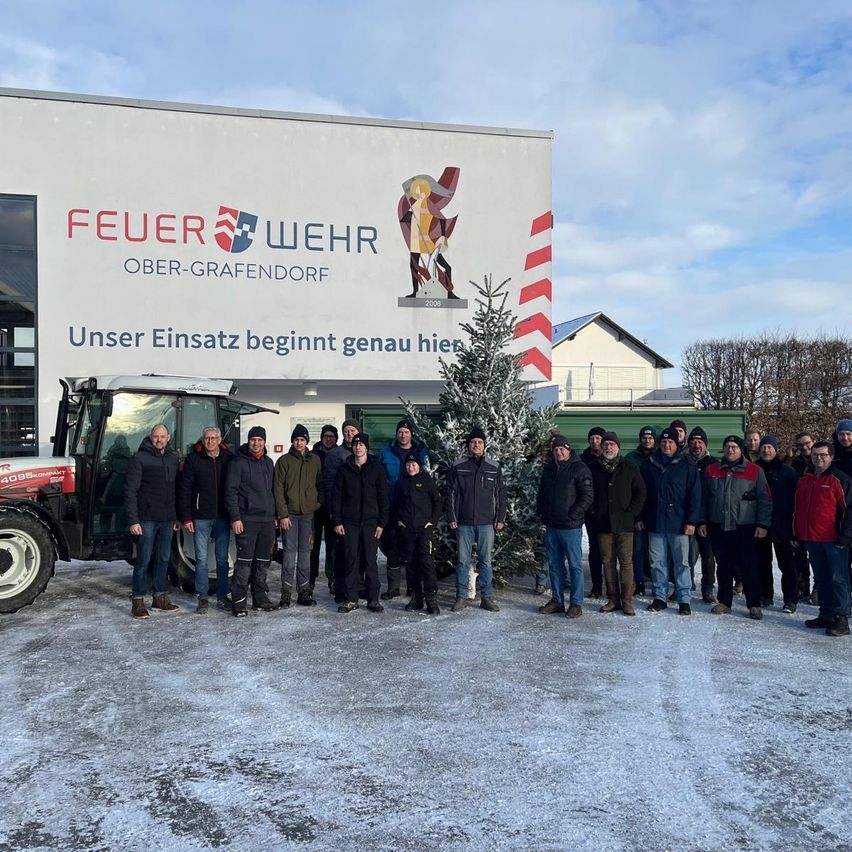 Eine Gruppe von Personen steht vor einem Gebäude mit einem Schild, das 'Feuer Wehr Ober-Grafendorf' anzeigt, was darauf hinweist, dass es sich um eine Feuerwehrstation handelt. Sie sind für kaltes Wetter warm gekleidet. Ein Traktor ist links neben der Gruppe geparkt.
