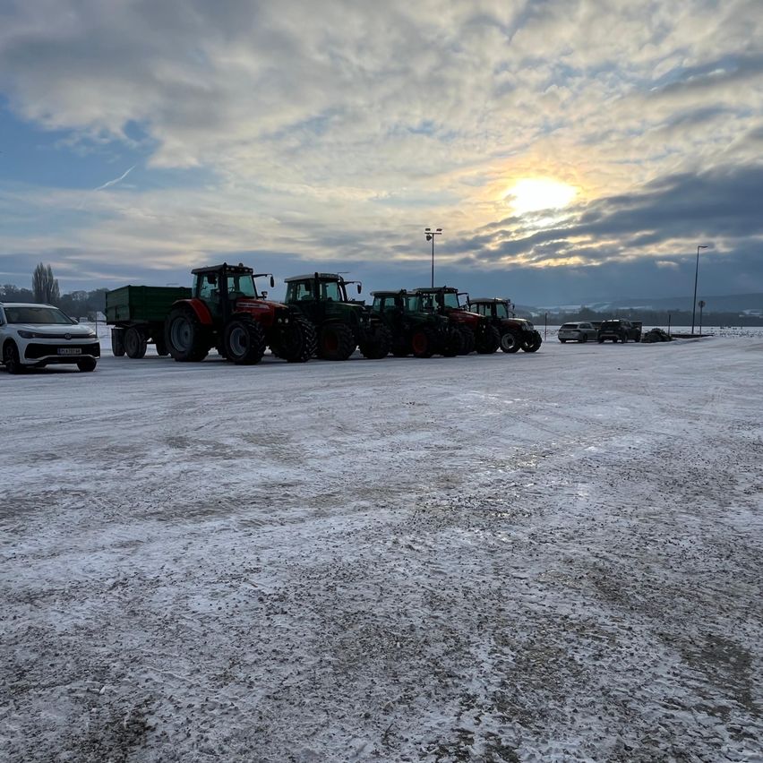 Mehrere Traktoren sind auf einem verschneiten Feld unter einem bewölkten Himmel geparkt. Einer von ihnen ist ein weißes Auto. In der Ferne gibt es Berge und Bäume.