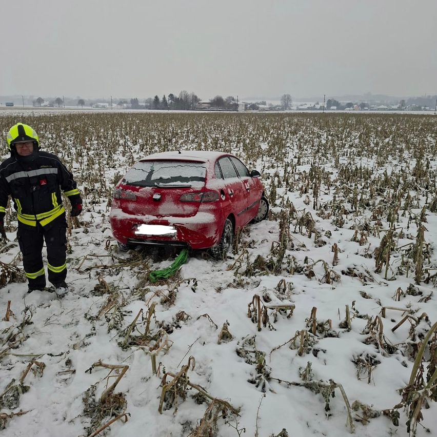 Ein Feuerwehrmann steht in einem verschneiten Feld neben einem roten Auto, das feststeckt. Das Auto ist mit Schnee bedeckt, und das Feld ist von kahlen Pflanzen umgeben.