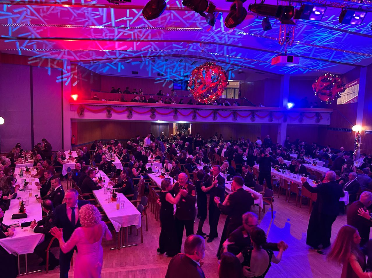 Ein großer Saal mit vielen Menschen in formeller Kleidung ist mit lila und roten Lichtern dekoriert. Paare tanzen auf der Tanzfläche, während andere an Tischen sitzen. Die Decke hat dekorative Lichter und einen Projektor.