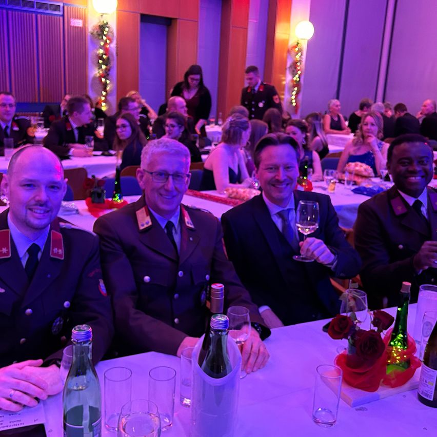 Eine Gruppe von Männern in Uniformen und Anzügen sitzt an einem Banketttisch, lächelt und hält Weingläser. Flaschen und Gläser stehen auf dem Tisch, und Leute sitzen hinter ihnen. Der Raum ist schwach beleuchtet mit lila Beleuchtung.