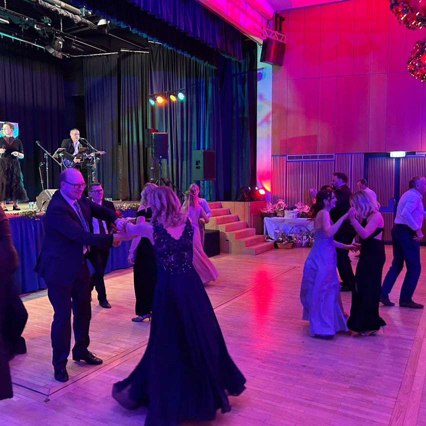 Eine Gruppe von Menschen tanzt auf einer Bühne in einem großen Saal, mit Musikern, die auf der Bühne spielen, und farbigen Lichtern, die den Bereich erhellen.