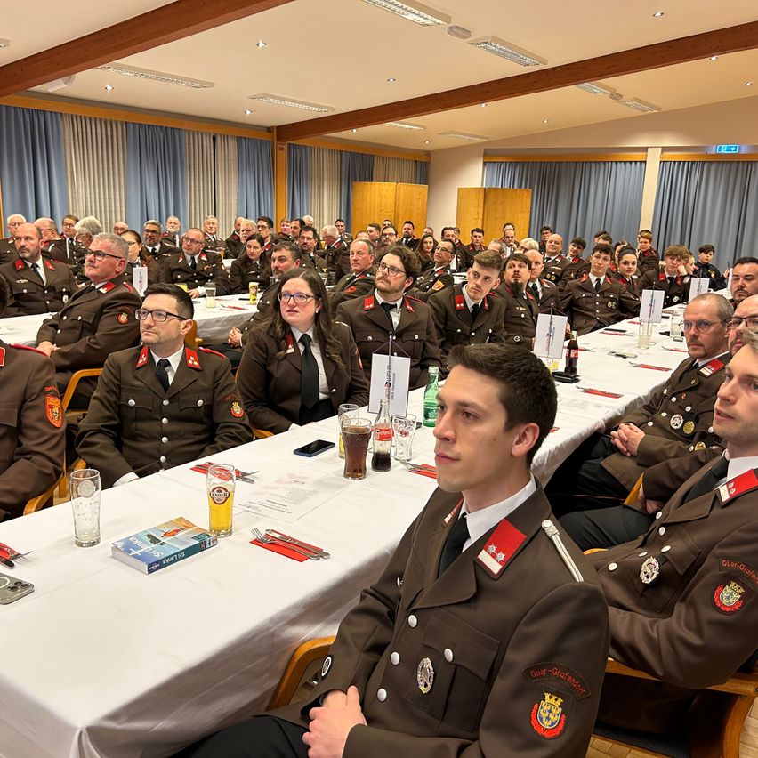 A large group of people in military uniforms sit around a long table. They are in a conference hall with blue curtains and overhead lights. There are glasses, bottles, and a book on the table.