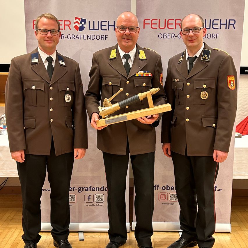 Three men in military uniforms stand before a backdrop with logos, holding a trophy. They are smiling and posing for a photo.