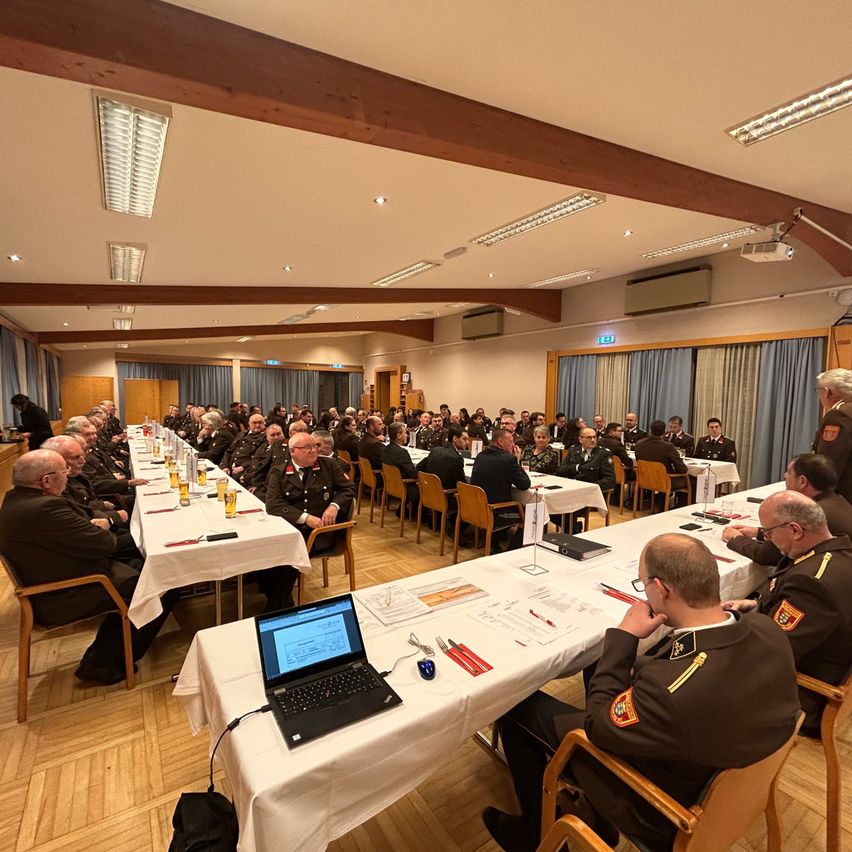 A conference room with military personnel in uniform seated at long tables, laptops and papers in front of them, a man in the background stands near a window with curtains.