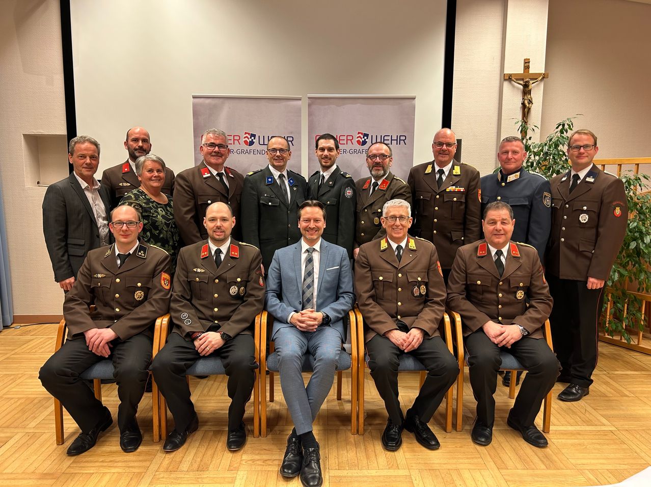 A group of men and women in uniforms are seated together in a conference room. A cross and a plant are visible in the background.