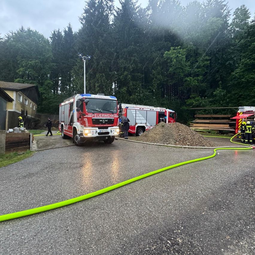 Ein Feuerwehrauto steht neben einem Haus. Ein grüner Schlauch liegt auf der Straße. Mehrere Feuerwehrleute sind um einen Haufen Kies herum.