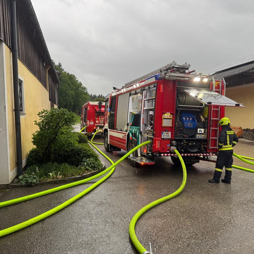 Ein Feuerwehrmann steht neben einem roten Feuerwehrwagen mit offenen Türen, grünen Schläuchen auf der Straße und einem weiteren Wagen im Hintergrund.