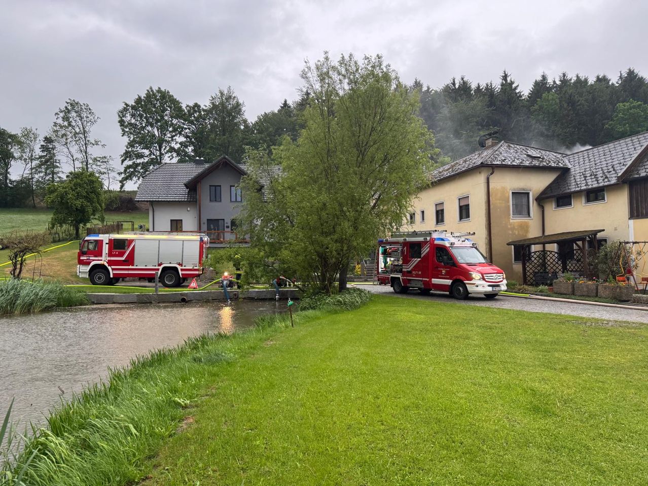 Zwei Feuerwehrwagen stehen auf der Straße in der Nähe eines Hauses mit Garten. Die Feuerwehrleute benutzen einen Schlauch im Wasser. Das Gebiet ist von Bäumen und Pflanzen umgeben.