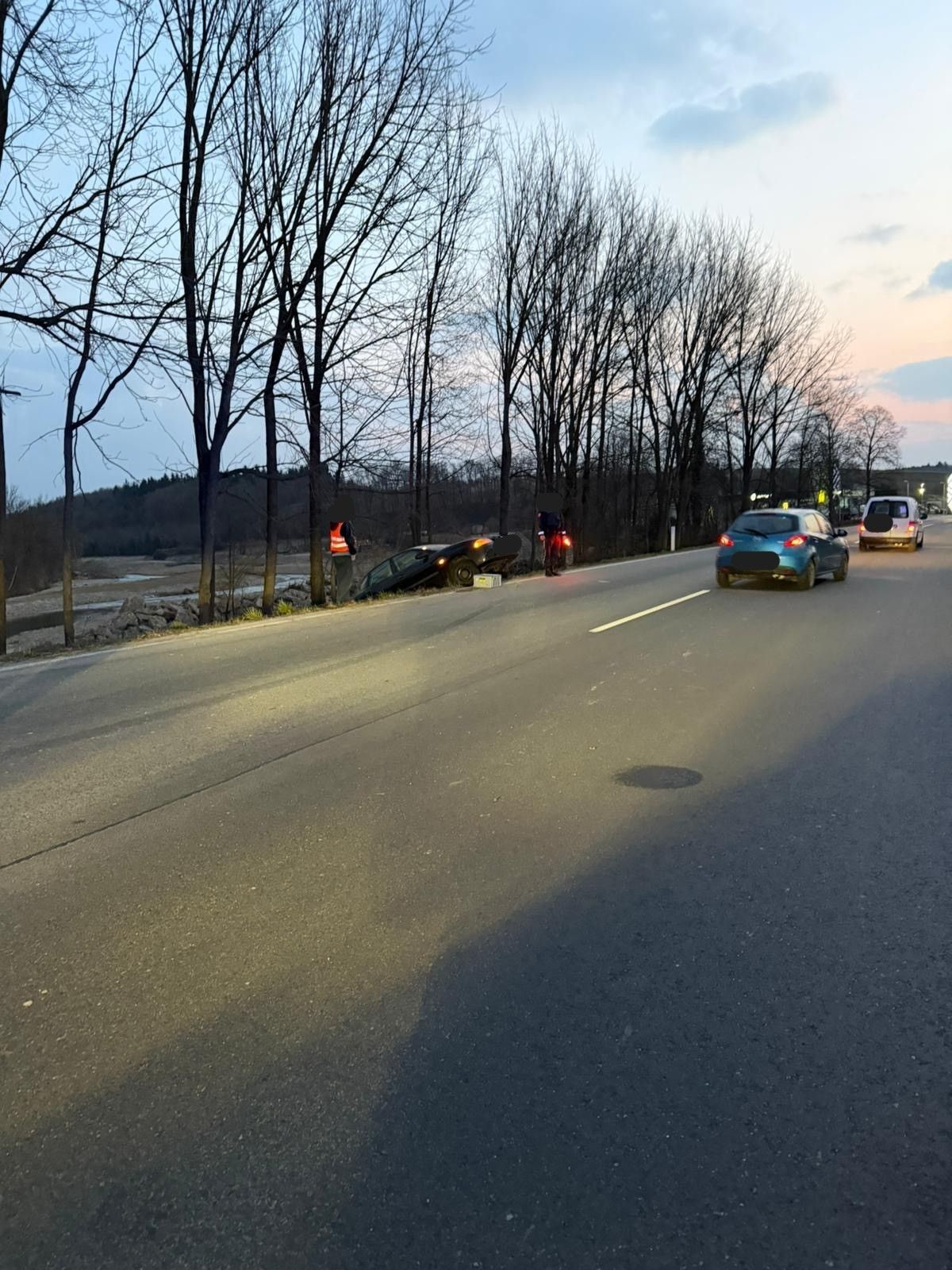 Ein Auto liegt auf der Seite der Straße, mit einer Person in der Nähe. Zwei Autos fahren auf der Straße vorbei.