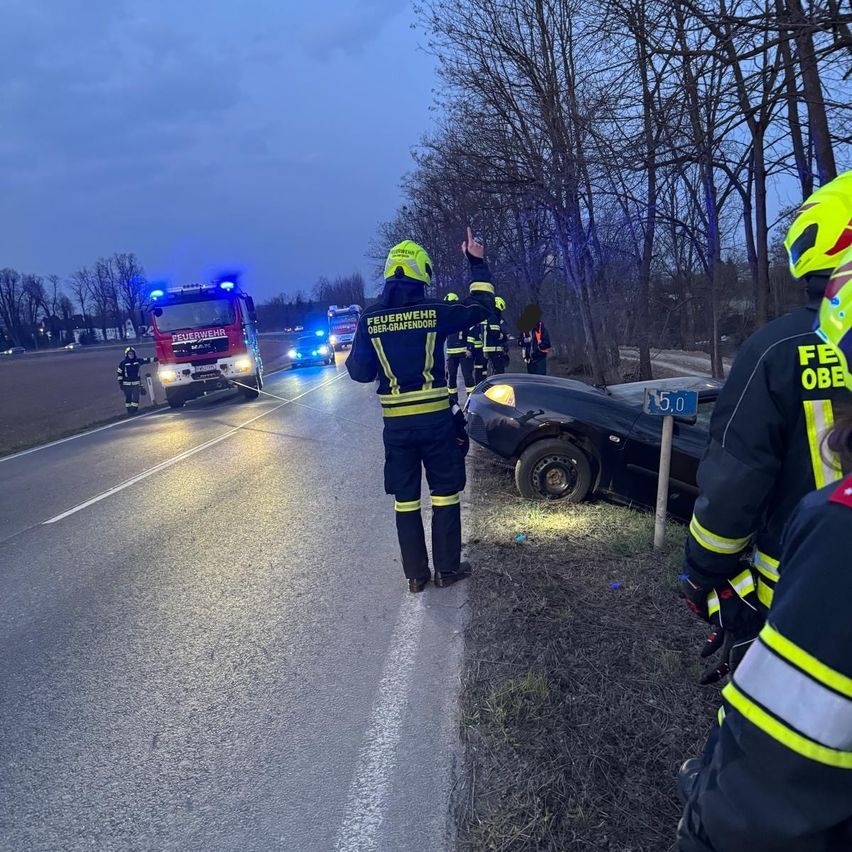 Rettungskräfte kümmern sich um einen Verkehrsunfall auf einer Straße, wobei ein Feuerwehrwagen und Polizeiautos anwesend sind.