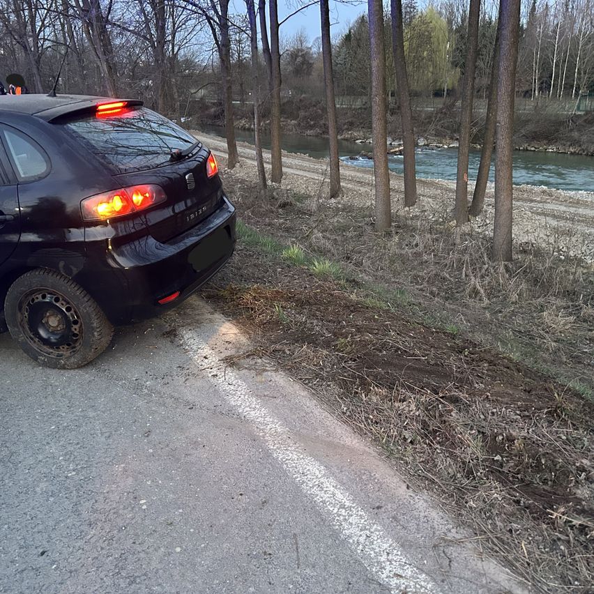Ein schwarzer Wagen ist am Straßenrand geparkt, nahe einem Fluss. Bäume stehen rechts, und eine weiße Linie ist auf der Straße.