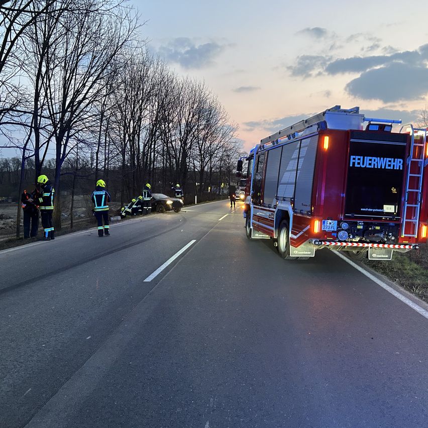 Ein roter Feuerwehrwagen mit dem Wort Feuerwehr auf der Rückseite ist am Straßenrand geparkt. Einige Menschen in gelben Westen und Helmen stehen am Straßenrand. Ein Auto ist kaputt und zwei Menschen stehen davor.