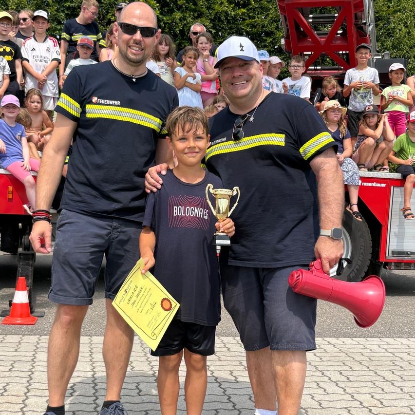 Ein junger Junge steht zwischen zwei Männern, hält einen Pokal und ein Zertifikat in der Hand, mit einem Feuerwehrwagen und einer Menge im Hintergrund.