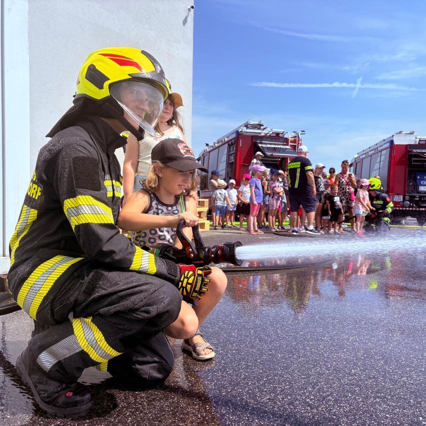 Ein Feuerwehrmann kniet auf nassem Asphalt und sprüht Wasser aus einem Schlauch, während ein junges Mädchen den Strahl hält. Dahinter beobachtet eine Menge Kinder und Erwachsene, mit geparkten Feuerwehrautos.