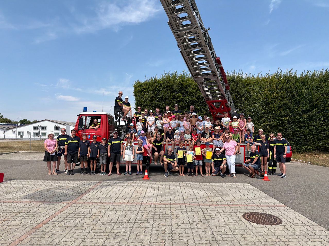 Eine Gruppe von Menschen, einschließlich Kindern, posiert für ein Foto vor einem Feuerwehrauto. Einige stehen auf dem LKW, während andere am Boden stehen.
