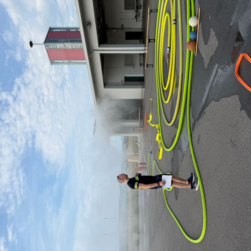 Ein Feuerwehrmann steht vor einem Gebäude mit Rauch, hält ein Papier und schaut auf den Boden. Es gibt grüne Schläuche auf dem Boden.