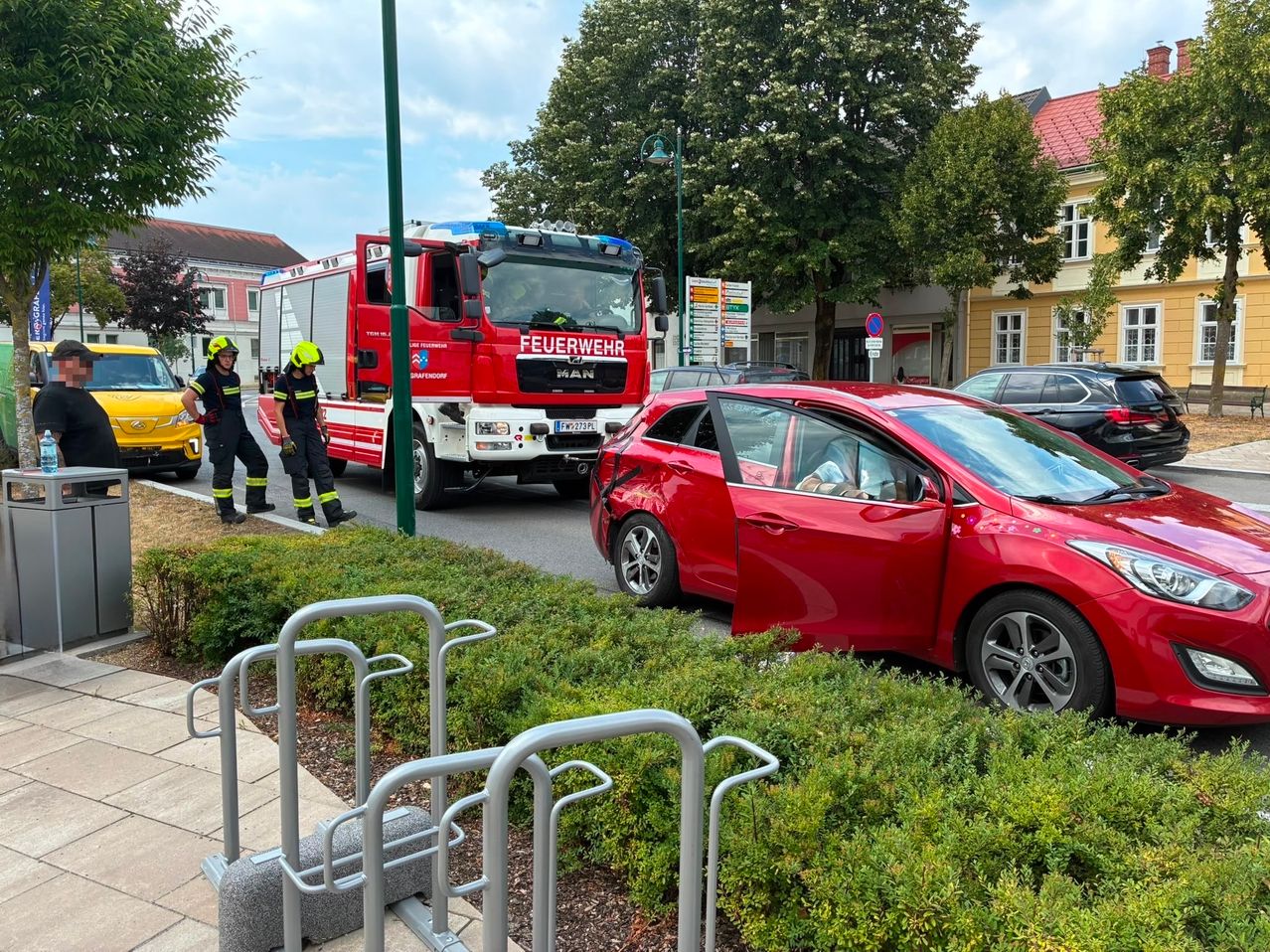 A red car is parked on the side of the road with its door open. Two firefighters are standing next to a fire truck. There are other cars parked nearby, and a streetlight is on the side of the road.