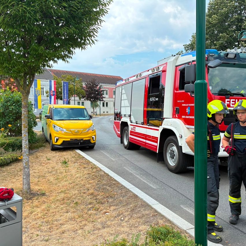 Two firefighters stand by a red fire truck on the side of the road, with a yellow car parked nearby. The scene is set in a town with a building and trees in the background.