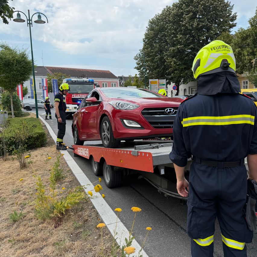 A red Hyundai car is being towed away by a tow truck, with a firefighter in a yellow helmet watching.