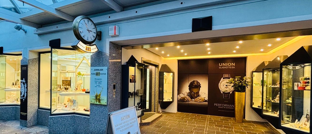 Eingangsbereich eines Juwelierladens mit einer Uhr und einem Schild, mit Glasfenstern, einem Ziegelboden und einem Poster eines Mannes mit einer Uhr.