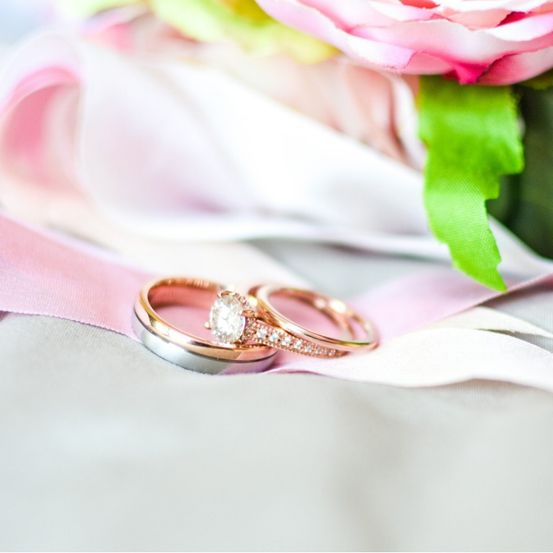Zwei goldene Eheringe mit Diamanten liegen auf einem rosa Stoff, mit einer rosa Rose im Hintergrund.