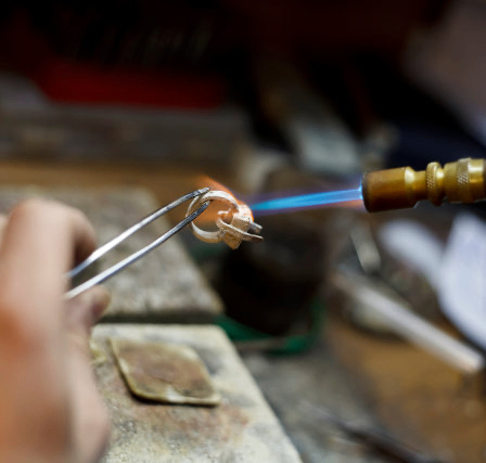 Eine Person benutzt eine Fackel, um einen mit einer Zange gehaltenen Ring zu erhitzen und konzentriert sich auf den Lötvorgang.