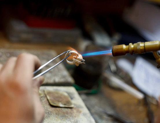 Eine Person benutzt eine Fackel, um einen mit einer Zange gehaltenen Ring zu erhitzen und konzentriert sich auf den Lötvorgang.