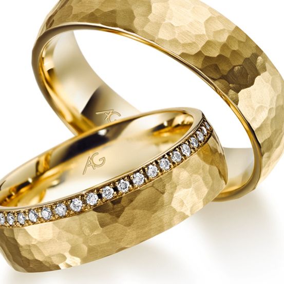 Zwei goldene Eheringe mit strukturierter Oberfläche und einer Reihe von Diamanten am Innenrand. Die Ringe liegen eng beieinander, möglicherweise für eine Hochzeitszeremonie.