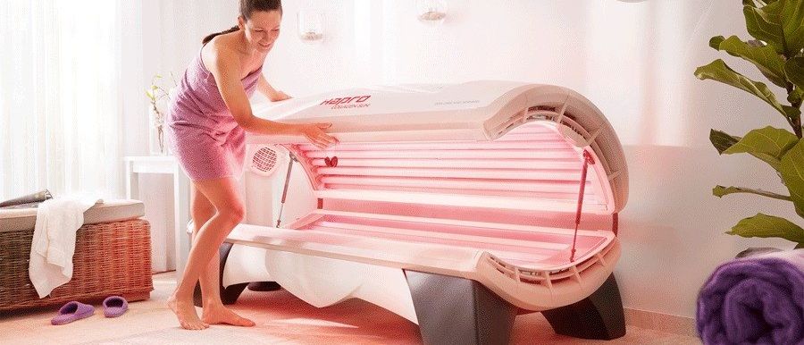 Eine Frau steht in einem Raum und bereitet sich darauf vor, ein rosa Solarium mit weißem Rahmen zu benutzen. Sie trägt ein lila Kleid.